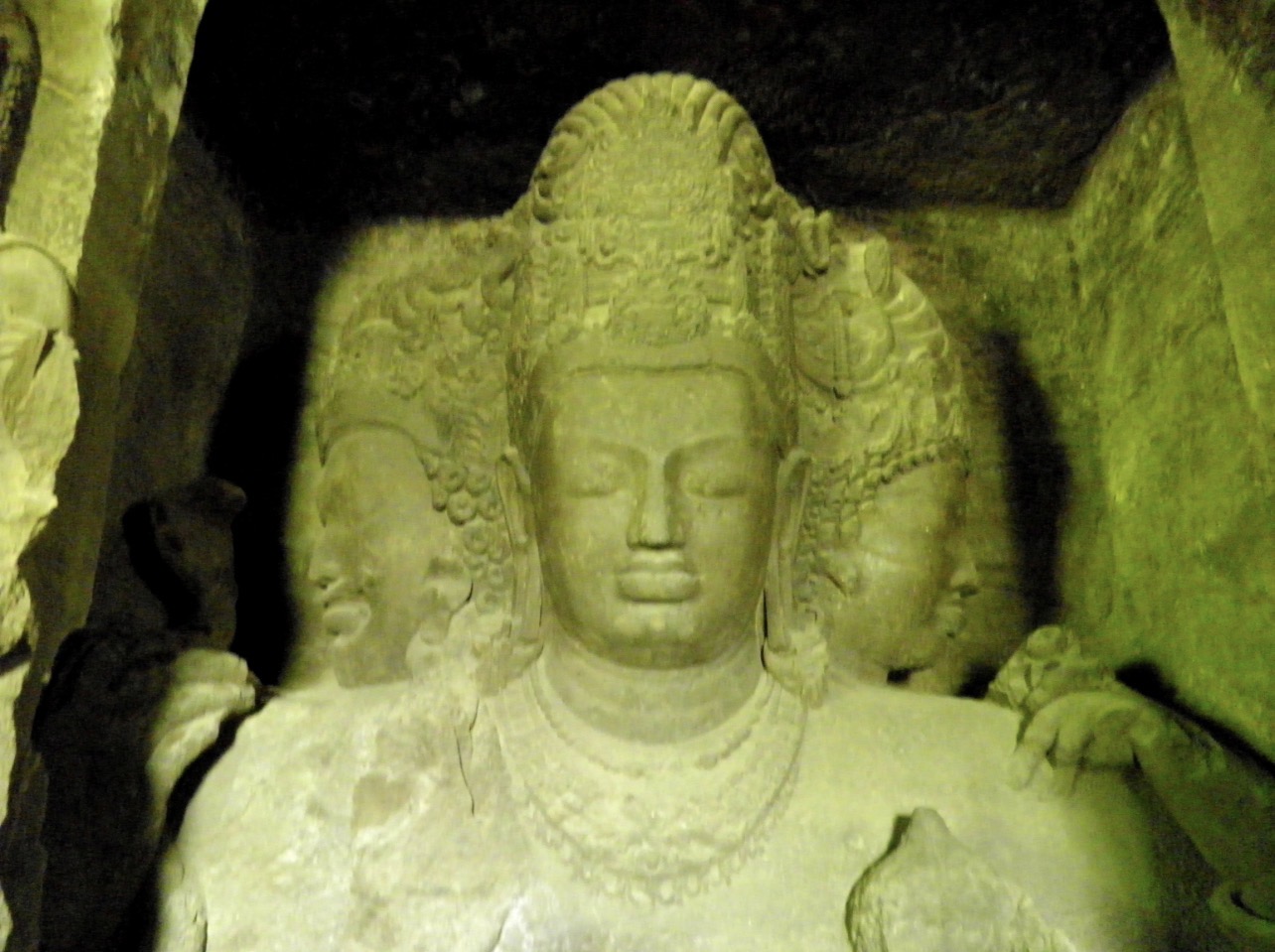 Ancient carved stone statue of Shiva lit by soft green light inside a cave temple
